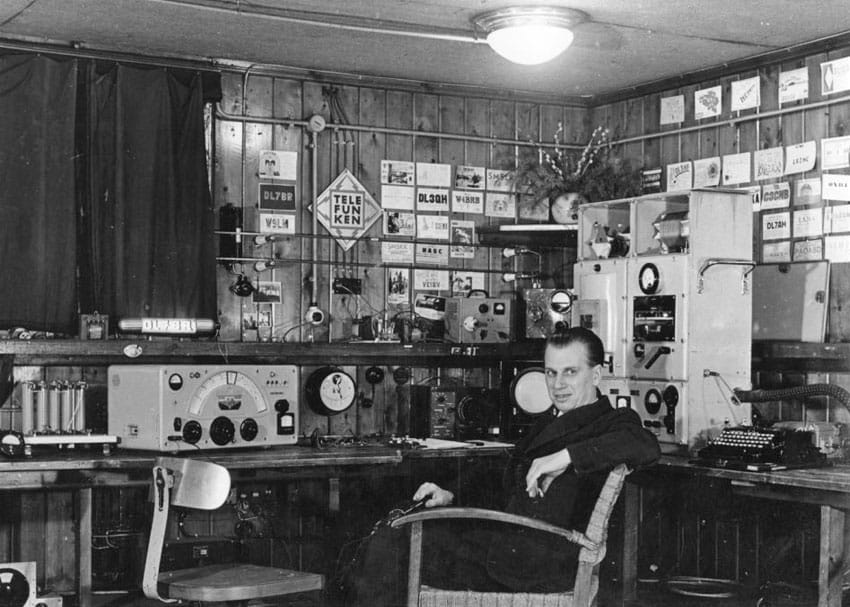 Well-dressed man sitting in front of old ham radio equipment with QSL cards on the wall